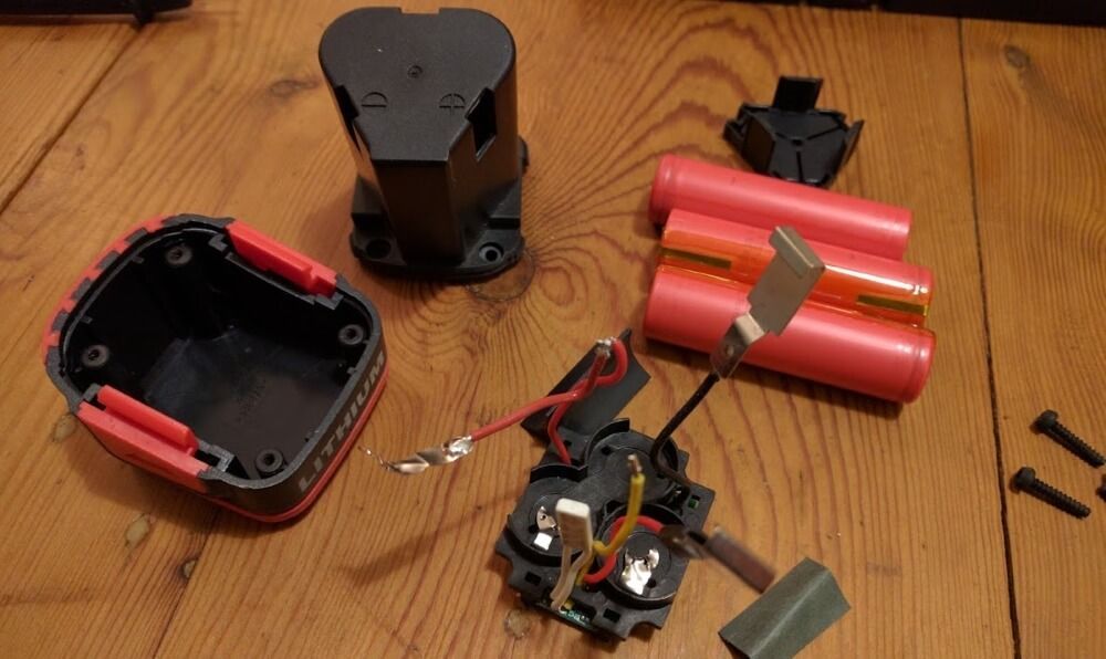 Bloc-batterie d'outil sans fil démonté, avec cellules internes au lithium roses, câblage et boîtier en plastique, sur un sol en bois.