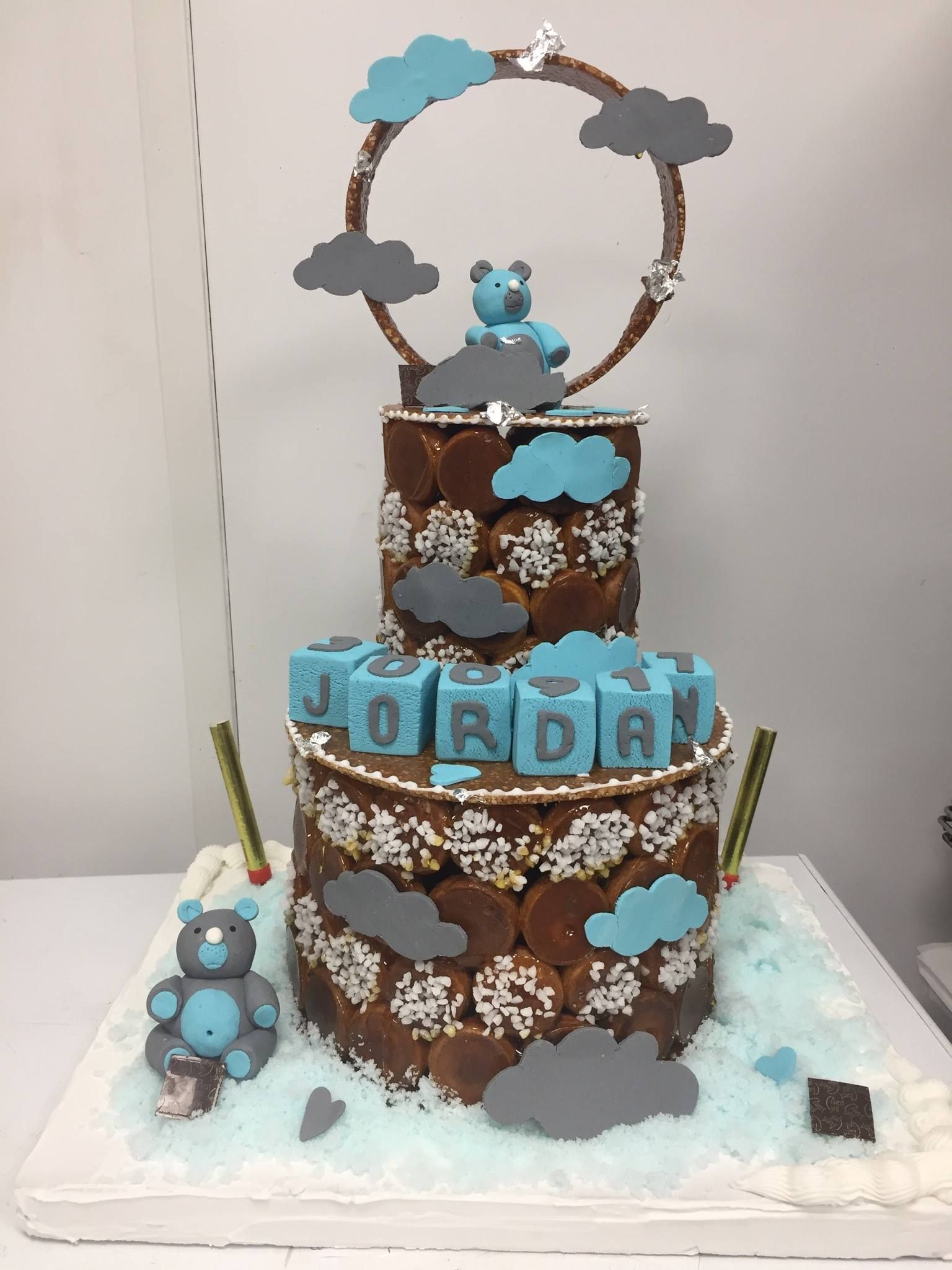 Un gâteau à deux étages de type profiterole, décoré de nuages bleus et gris et de figurines d'ours en peluche, portant le nom JORDAN.