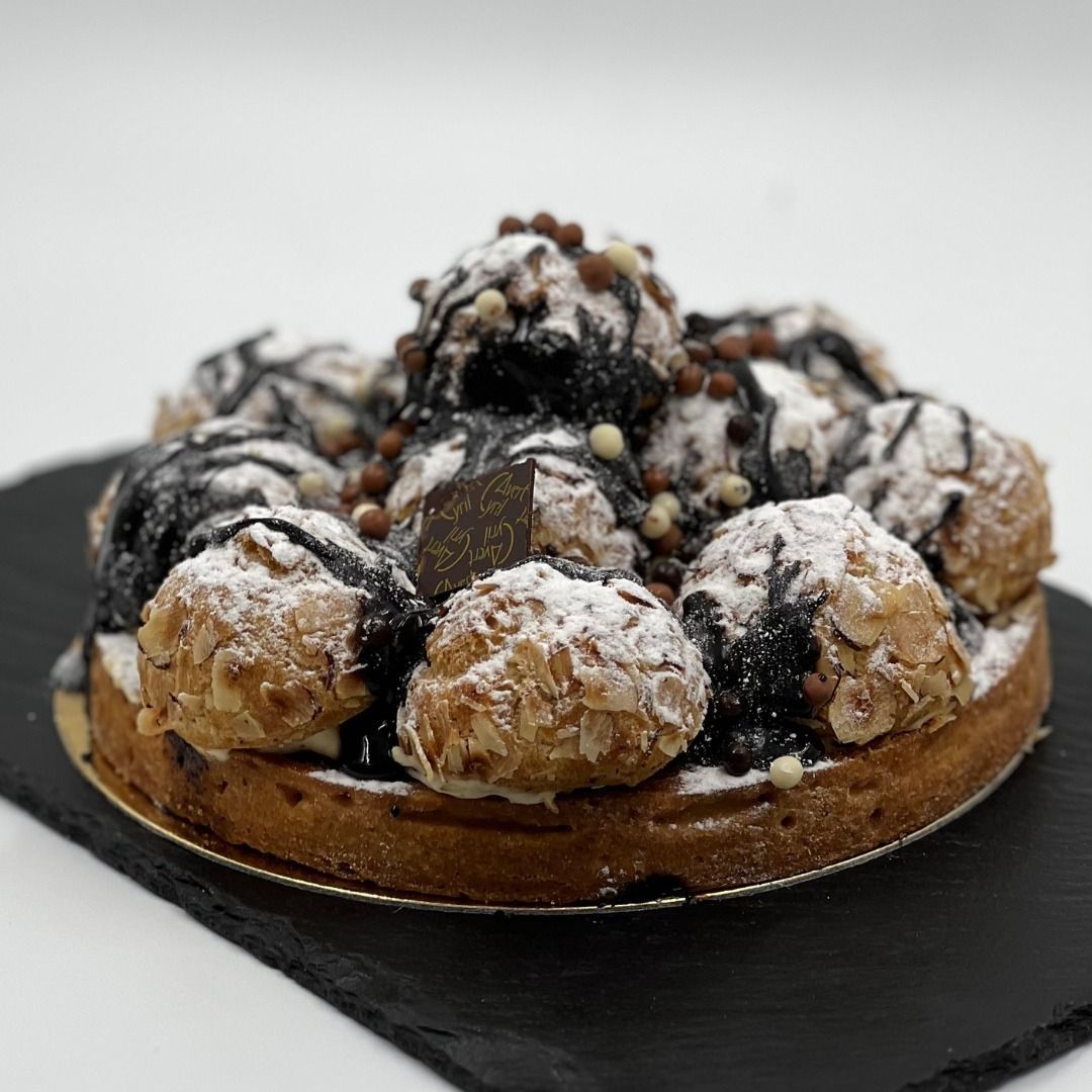 Une pâtisserie Saint-Honoré composée de choux fourrés à la crème, d'un filet de chocolat et de sucre glace, présentée sur un plateau de service en pierre foncée.