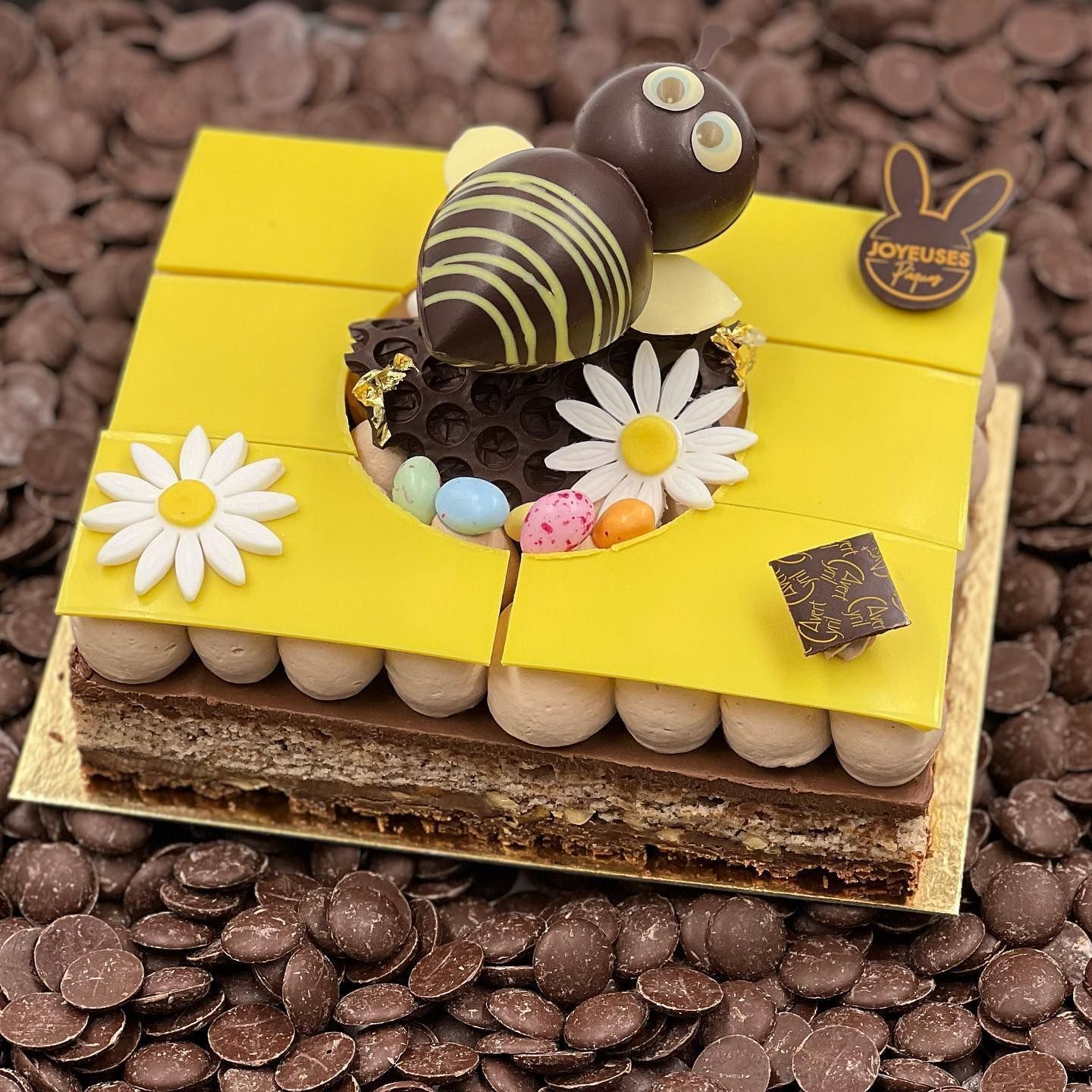 Un gâteau au chocolat carré à plusieurs étages, garni d'une abeille en chocolat, de marguerites blanches et de petits œufs colorés sur des pépites de chocolat.