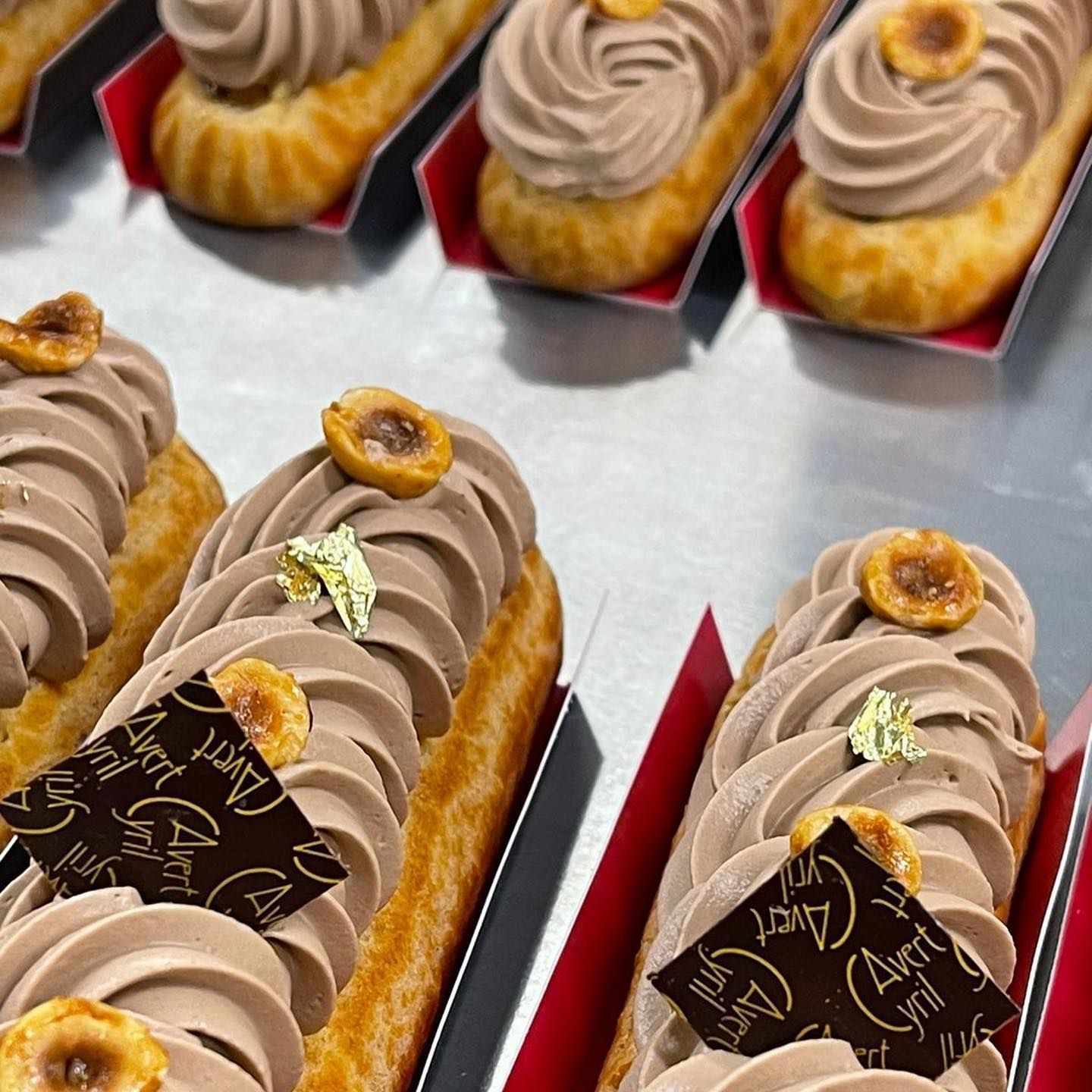 Des rangées d'éclairs aux noisettes, nappés de glaçage au chocolat, de feuilles d'or et de noisettes grillées, présentés dans des plateaux rouges individuels.