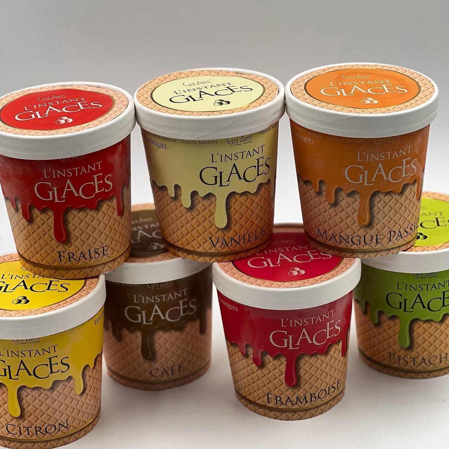 Sept pots de crème glacée L'Instant Glaces à bords gaufrés, disposés sur une surface blanche.