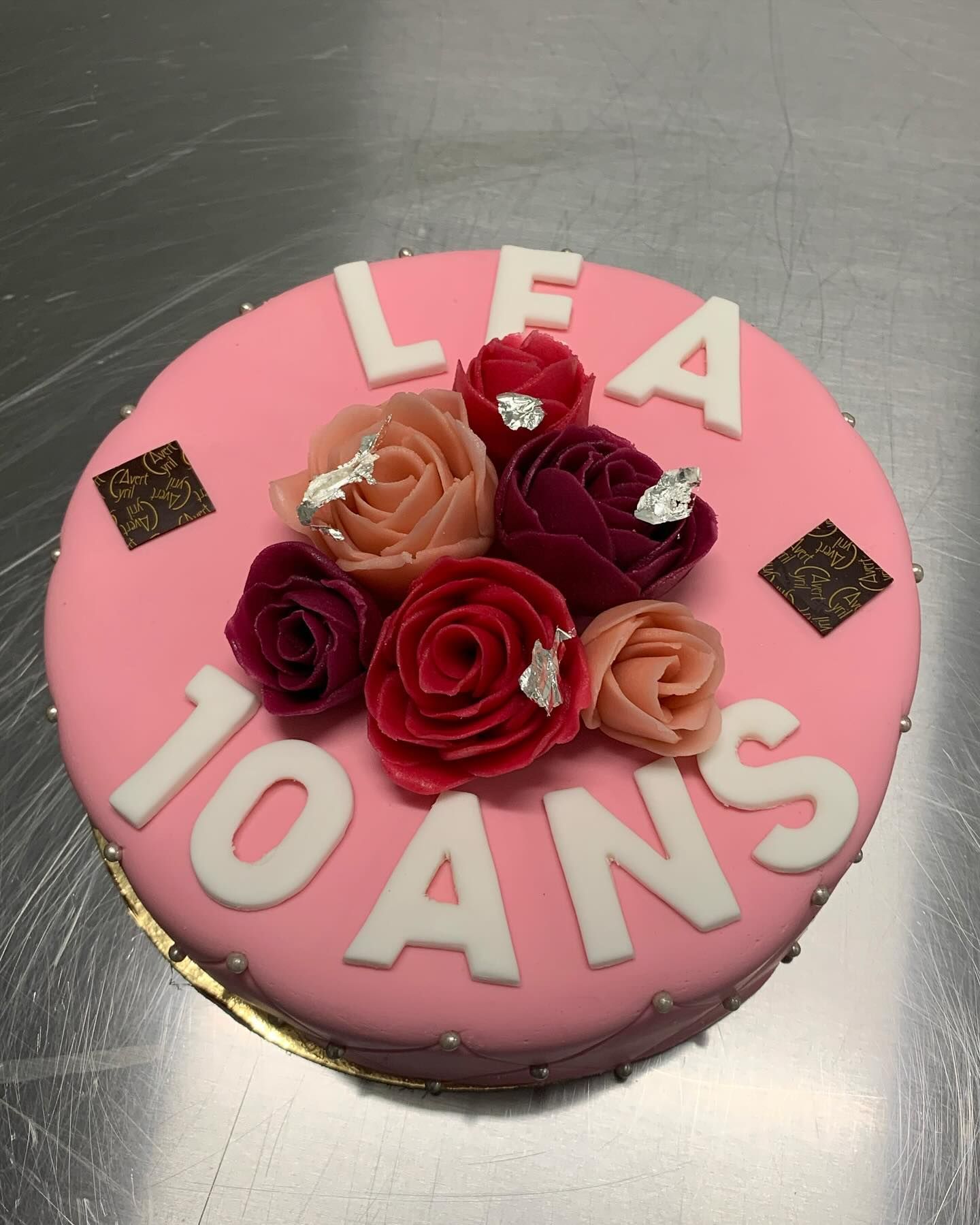 Un gâteau rond rose décoré de roses en glaçage coloré, de touches argentées et de l'inscription blanc LEA 10 ANS.