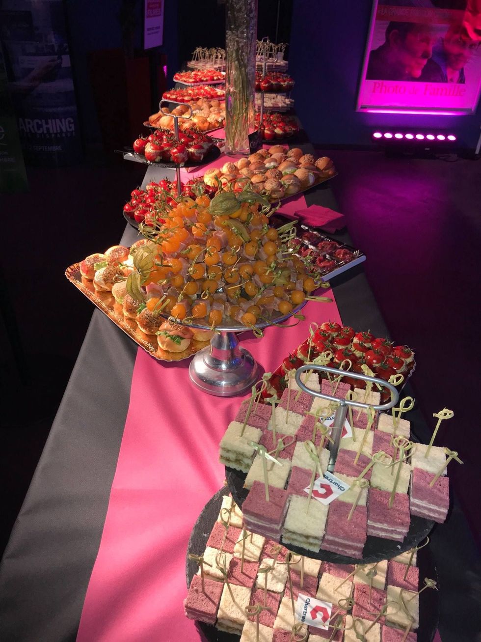 Une table de buffet recouverte d'un long chemin de table rose présente différents niveaux de petits amuse-bouche décoratifs, d'entrées et de sandwichs.
