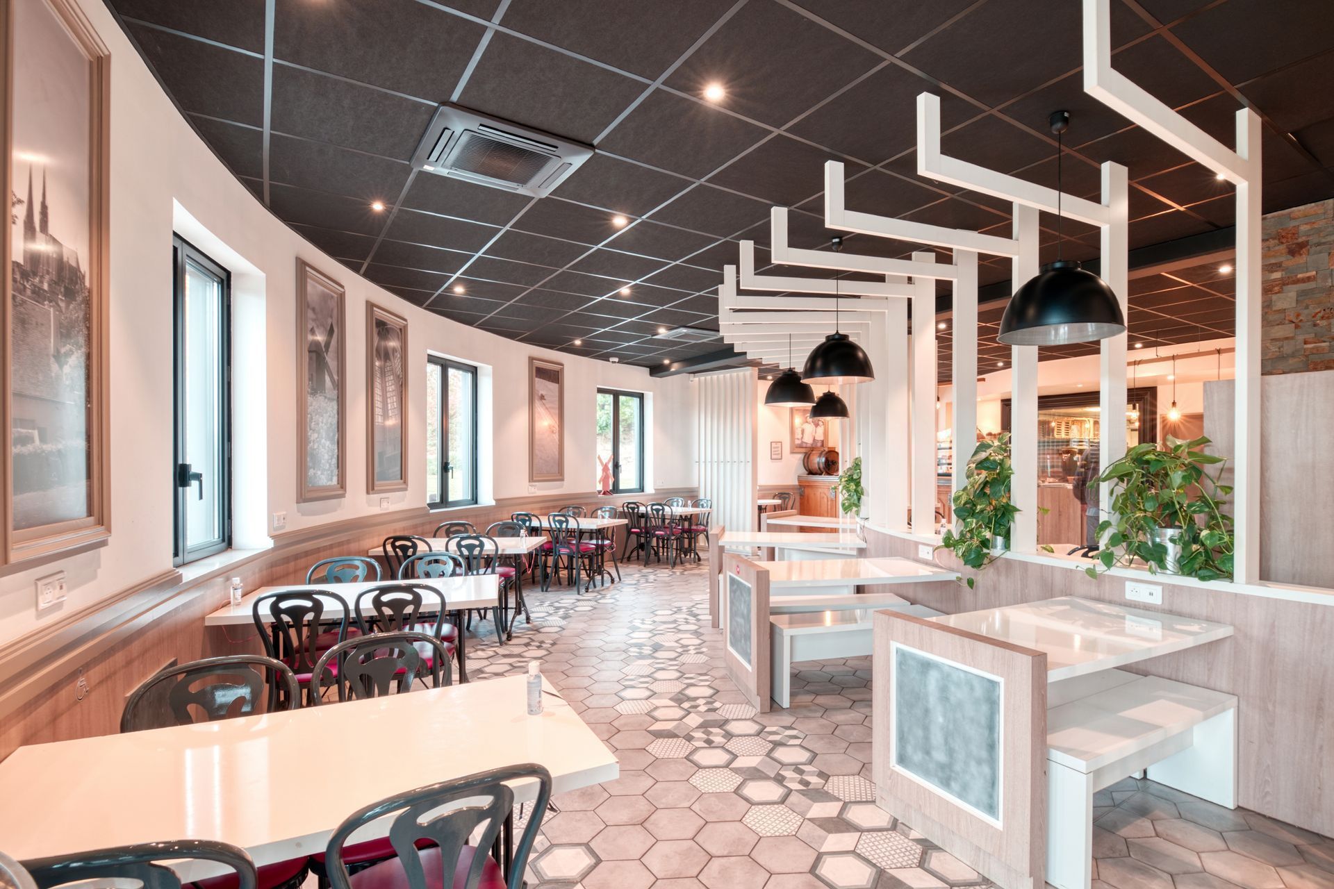 Vue intérieure d'un restaurant avec des tables rondes, des chaises noires, un plafond à motifs sombres et des poutres blanches décoratives.