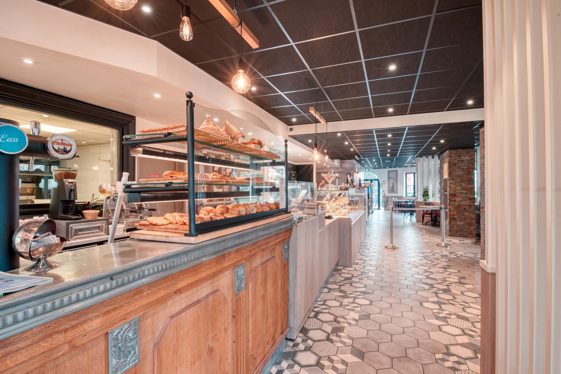 Intérieur d'une boulangerie avec un comptoir de service en bois, des étagères vitrées remplies de pâtisseries et un sol carrelé.