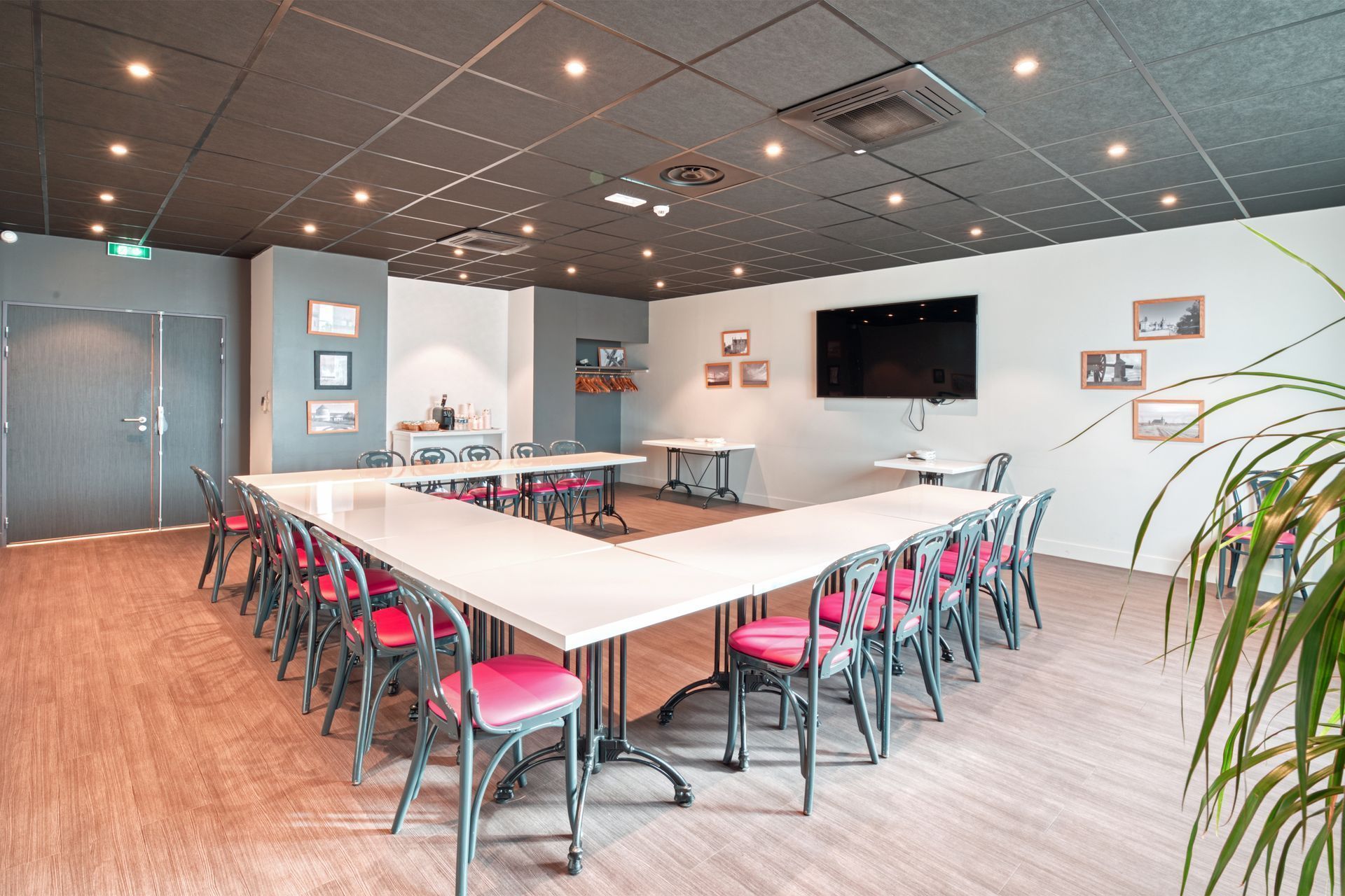 Table de conférence en forme de U avec des chaises roses dans une salle de réunion dotée d'un plafond en dalles acoustiques noires et d'une télévision murale.