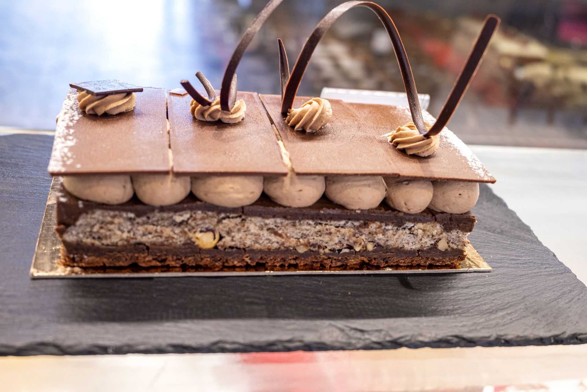 Un gâteau rectangulaire au chocolat avec des couches de noix, garni de volutes de crème, de rectangles de chocolat et de spirales décoratives.