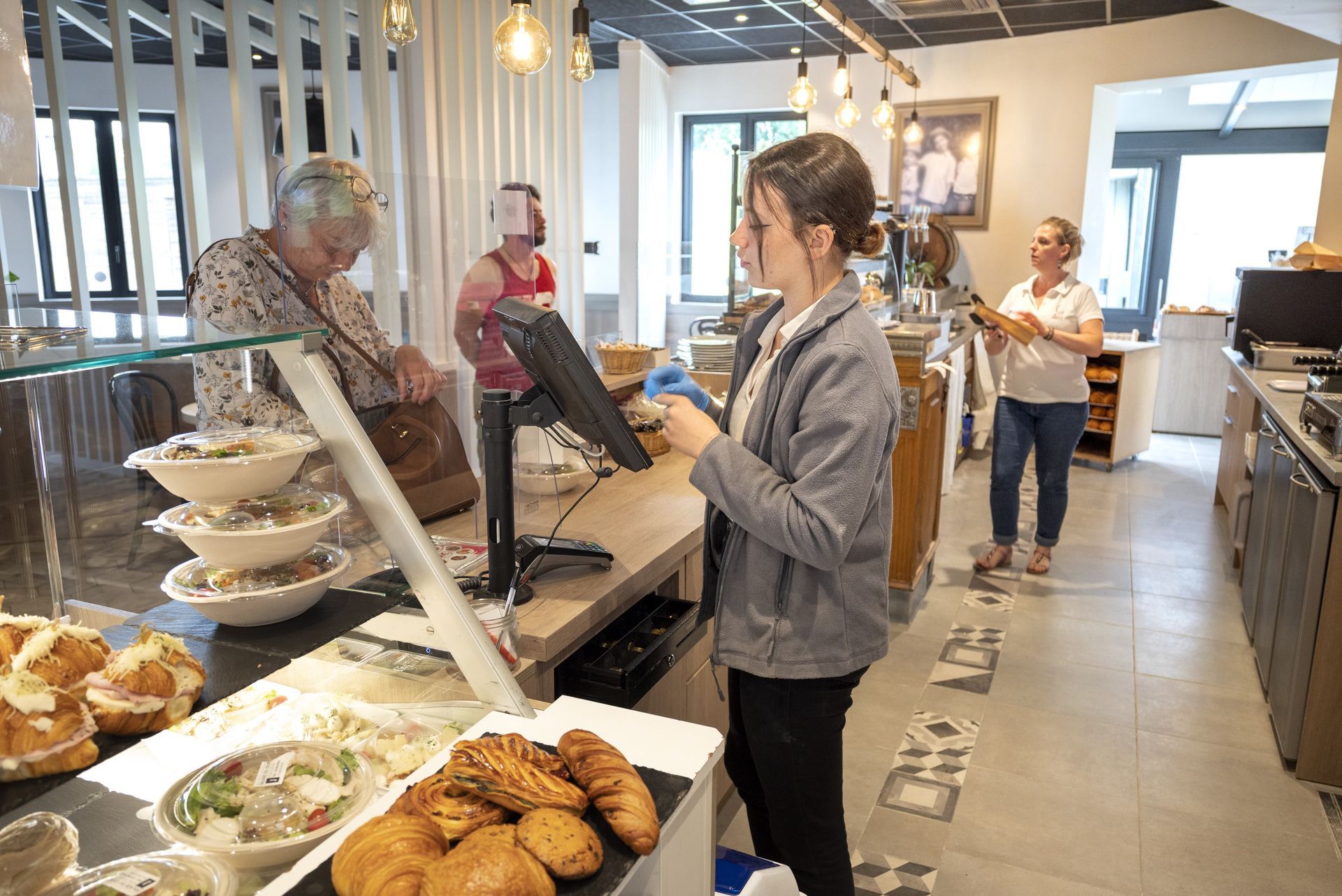 Un client de boulangerie discute avec un caissier au comptoir, tandis que d'autres font la queue au milieu des étagères remplies de produits de boulangerie.