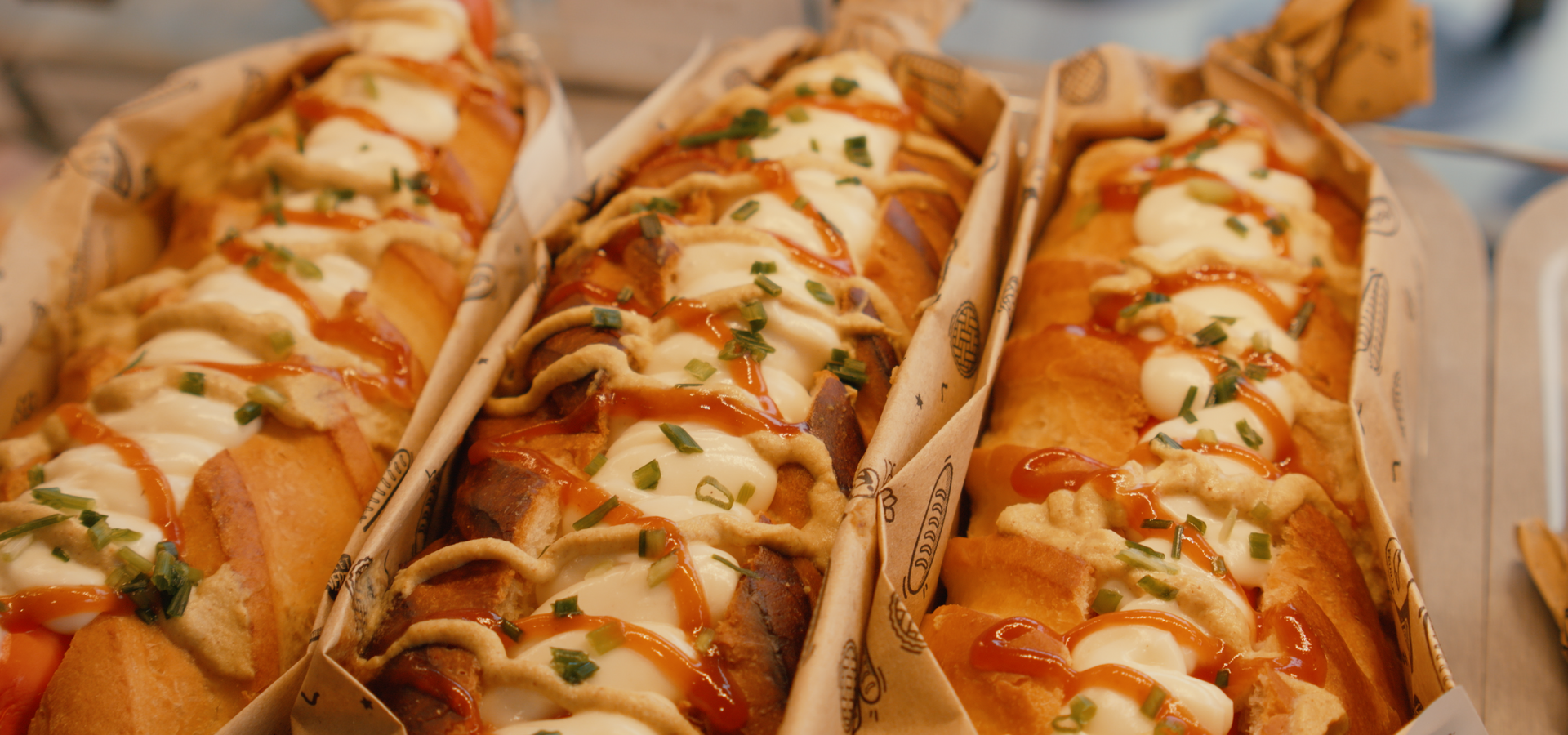 Trois longs sandwichs enveloppés dans du papier, garnis de volutes de sauce blanche, de sauce à l'orange et d'herbes vertes hachées.