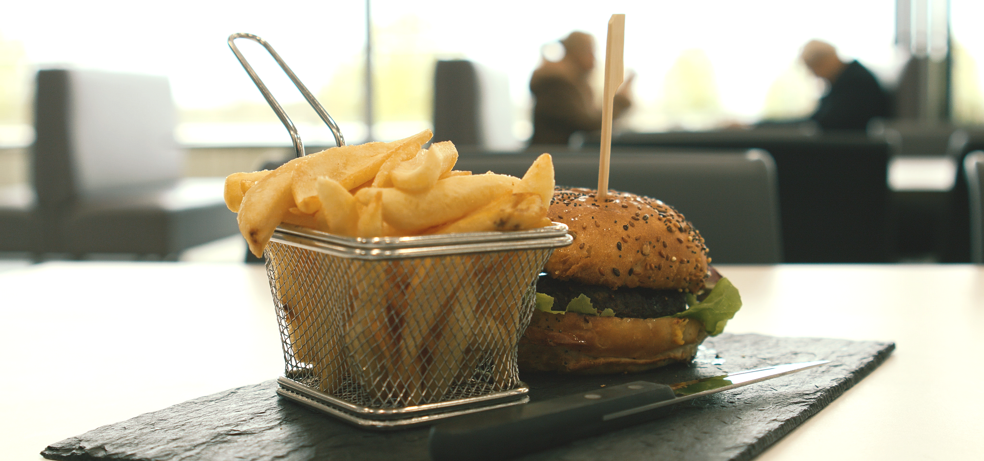Un hamburger et une corbeille de frites servis sur un plateau rectangulaire en ardoise sombre, dans un restaurant.