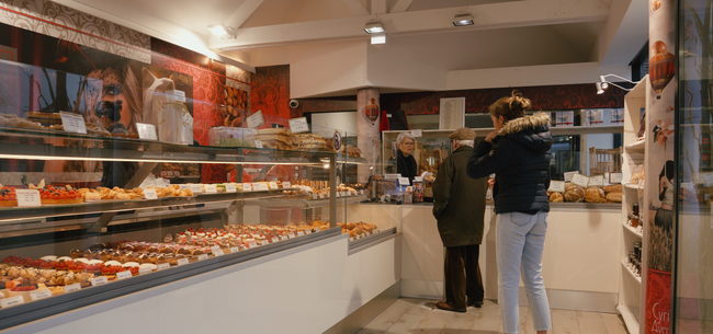 L'intérieur d'une boulangerie avec une vitrine remplie de pâtisseries et deux clients debout au comptoir.