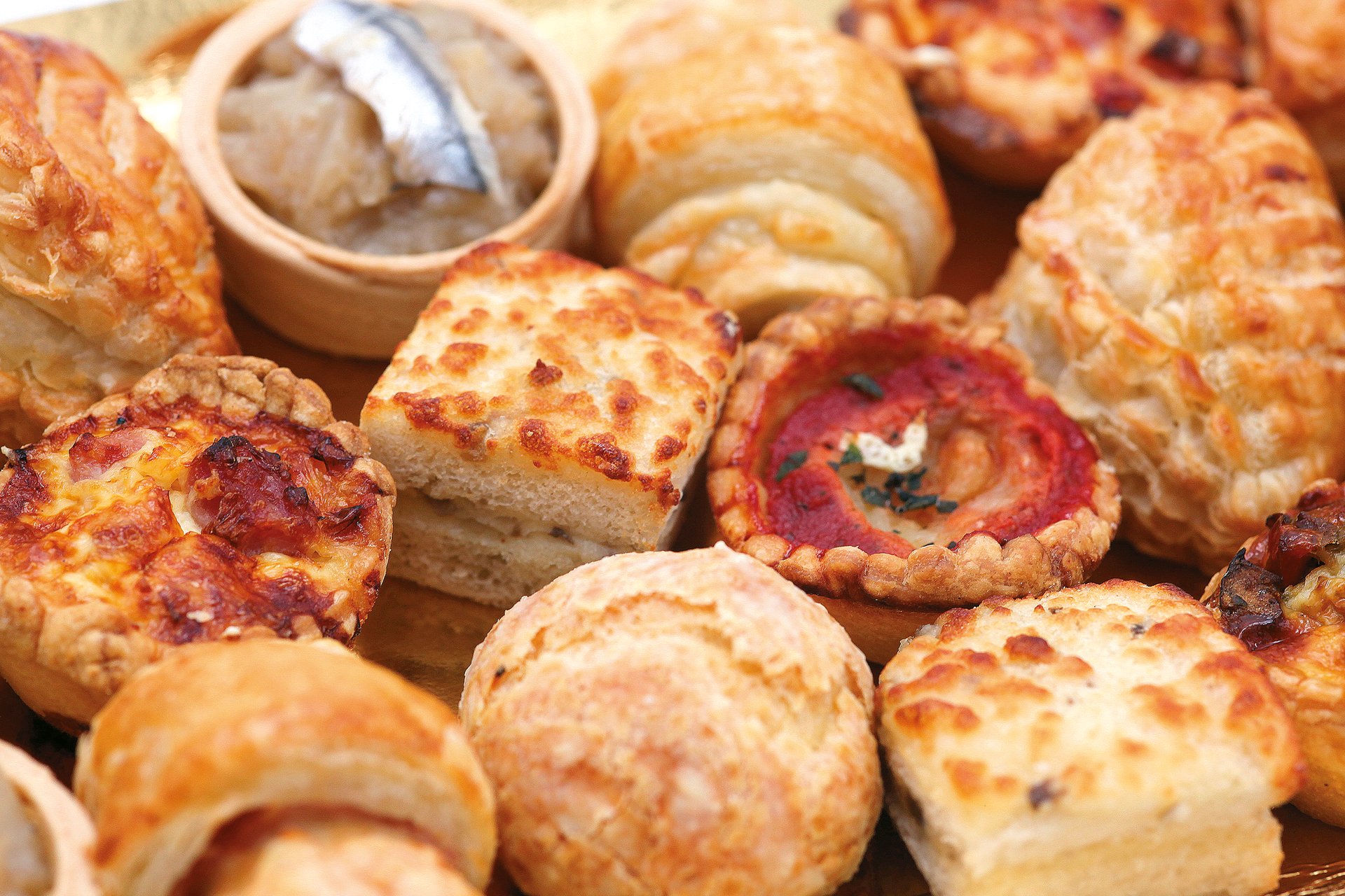 Un assortiment de pâtisseries salées dorées, de quiches et un petit bol de tartinade au poisson, disposés ensemble.