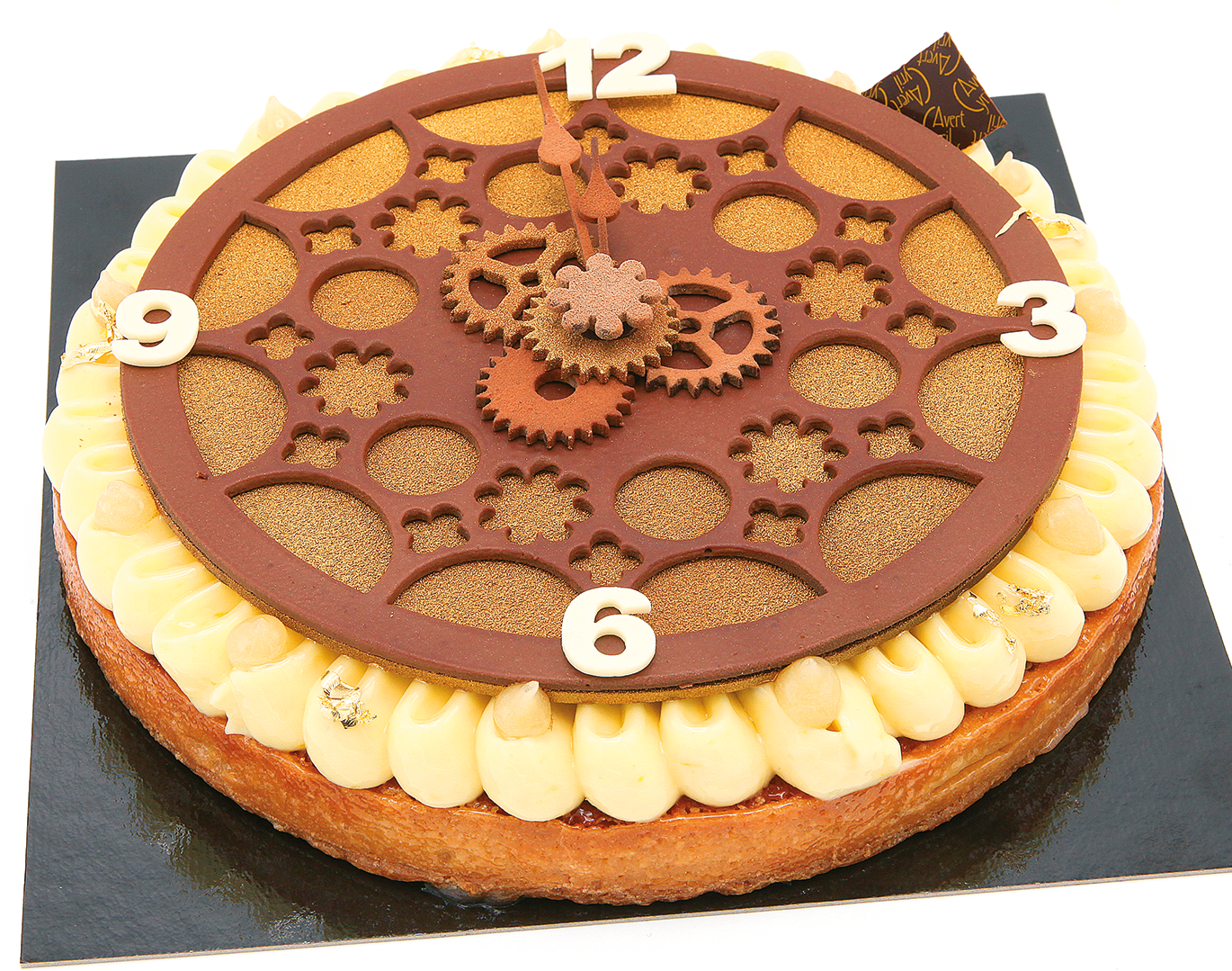 Une tartelette ronde décorée d'un motif d'engrenage d'horloge en chocolat brun, de quenelles de crème blanche et de chiffres blancs.