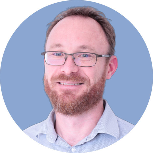 Homme souriant avec des lunettes et une barbe, sur fond bleu clair.