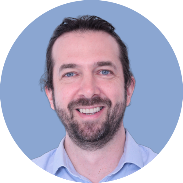 Un homme aux yeux bleus et à la barbe sourit, vêtu d'une chemise bleu clair, sur un fond circulaire bleu clair.