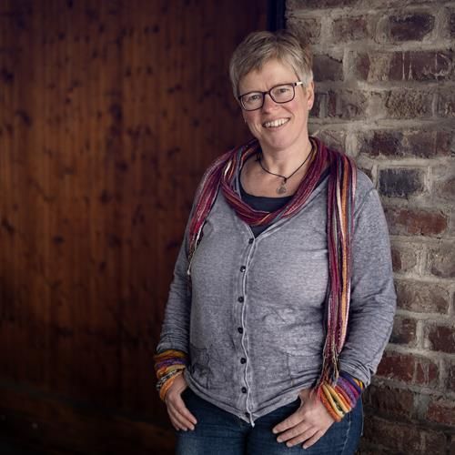 Eine Frau mit Brille und Kopftuch steht vor einer Backsteinmauer.