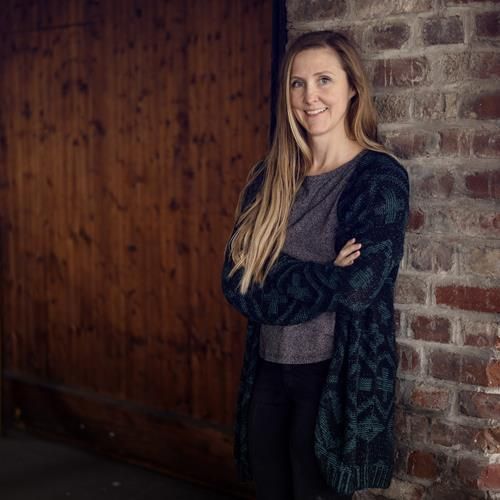 Eine Frau lehnt mit verschränkten Armen an einer Backsteinmauer und lächelt.