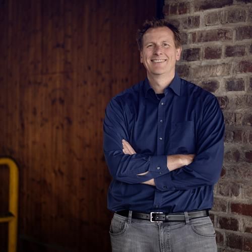 Ein Mann in einem blauen Hemd steht mit verschränkten Armen vor einer Backsteinmauer