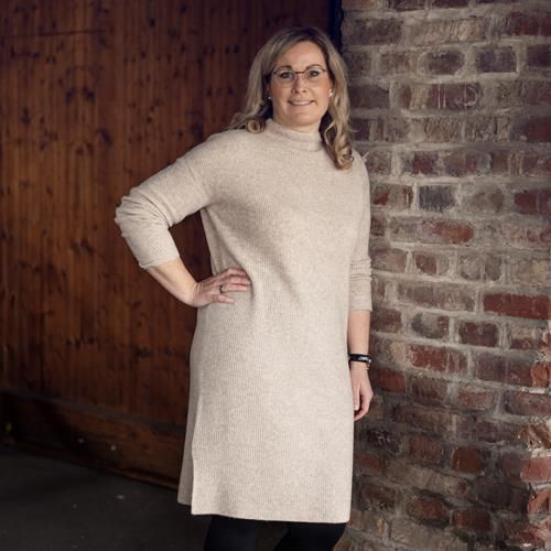 Eine Frau in einem beigen Kleid steht vor einer Backsteinmauer.