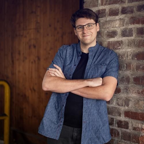 Ein Mann mit Brille und blauem Hemd steht mit verschränkten Armen vor einer Backsteinmauer.