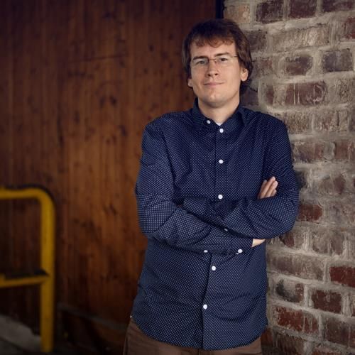 Ein Mann in einem blauen Hemd lehnt mit verschränkten Armen an einer Backsteinmauer.