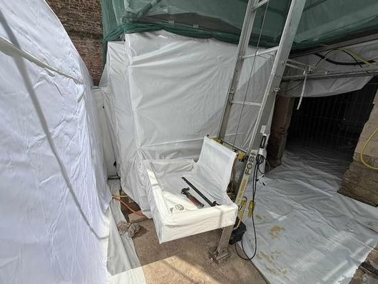 Chantier de construction avec une structure recouverte de blanc, un élévateur avec des outils et une bâche en plastique au sol.