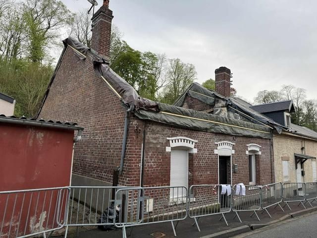 Maison en briques au toit effondré, jonchée de débris et entourée d'une clôture de protection, en bordure de rue.