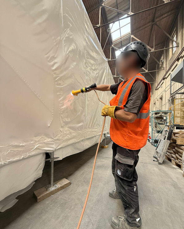 Une personne utilise un pistolet thermique sur une bâche en plastique dans un entrepôt, portant un équipement de sécurité.