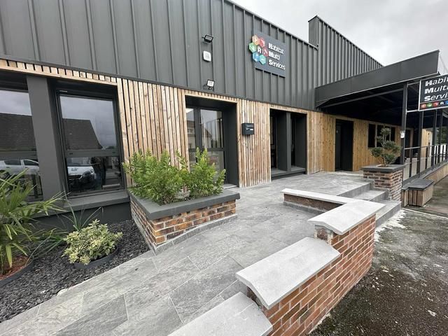 Vue extérieure d'un bâtiment commercial avec un revêtement en bois et gris, comprenant un petit parterre de jardin et un chemin d'accès.