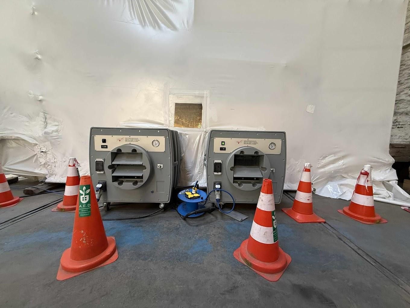 Deux machines grises entourées de cônes de signalisation, dans une zone aux murs blancs.