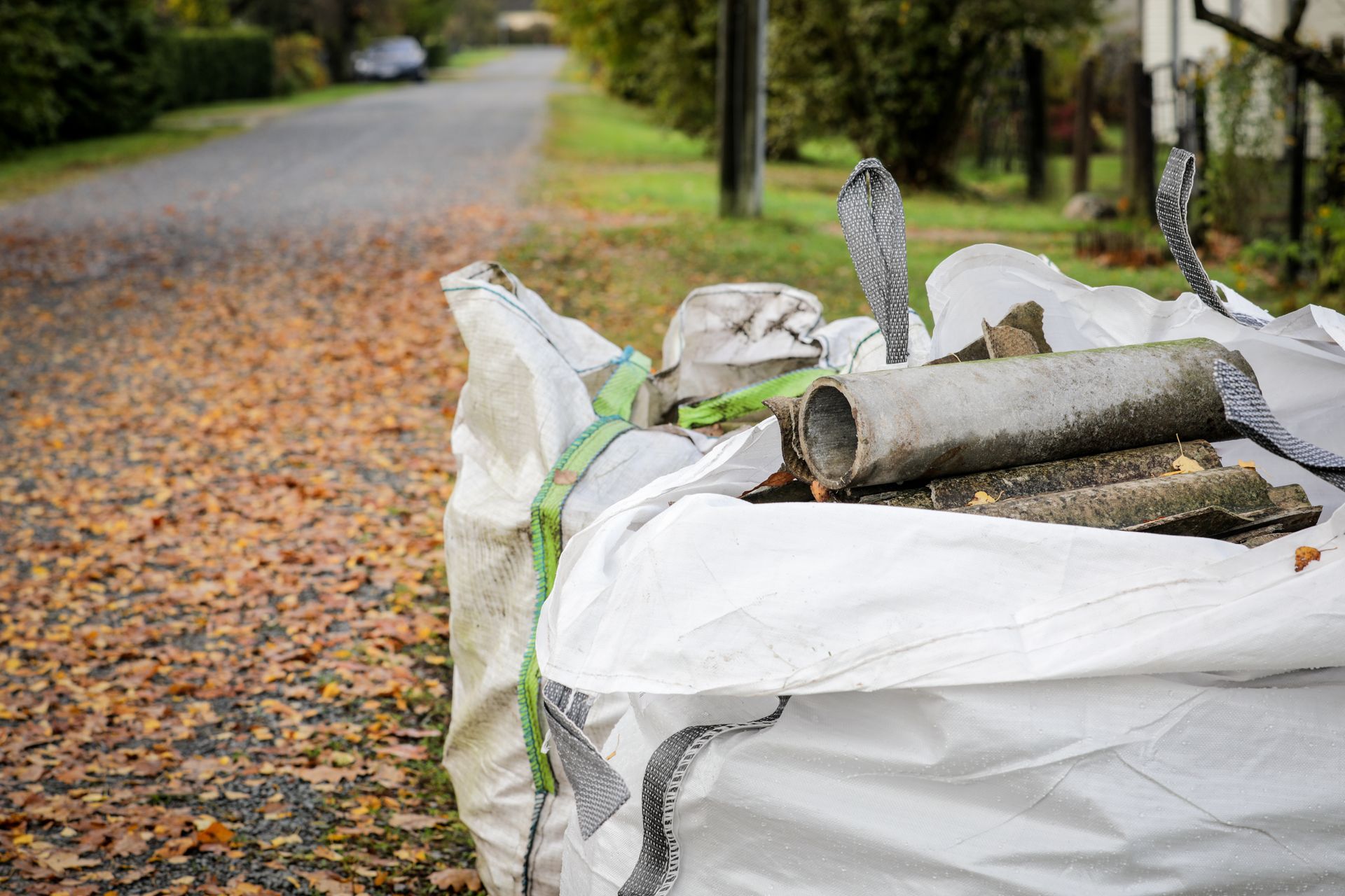 Un sac industriel blanc rempli de débris est posé au bord d'une route recouverte de feuilles d'automne.