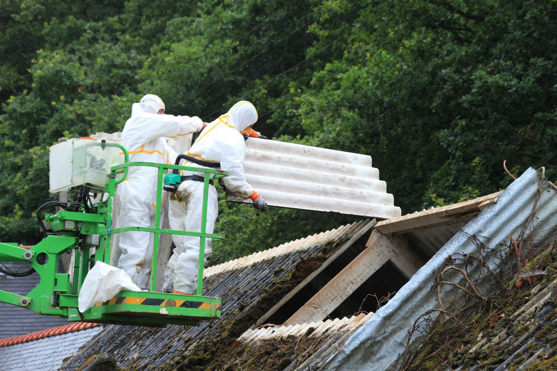 Des ouvriers en combinaisons de protection blanches retirent la toiture en amiante d'un bâtiment à l'aide d'une nacelle élévatrice.