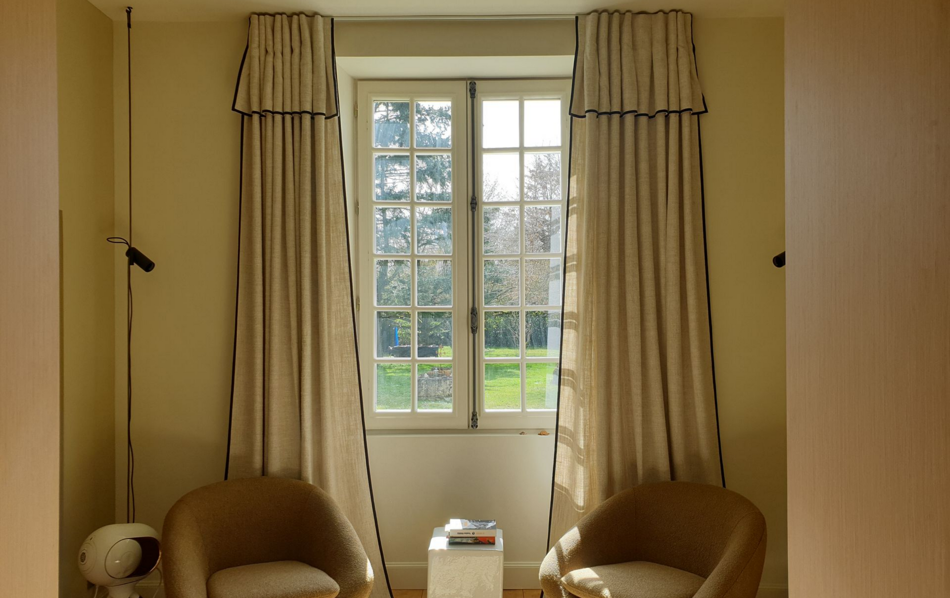 Fenêtre avec de longs rideaux beiges bordés de noir et de deux fauteuils couleur sable.