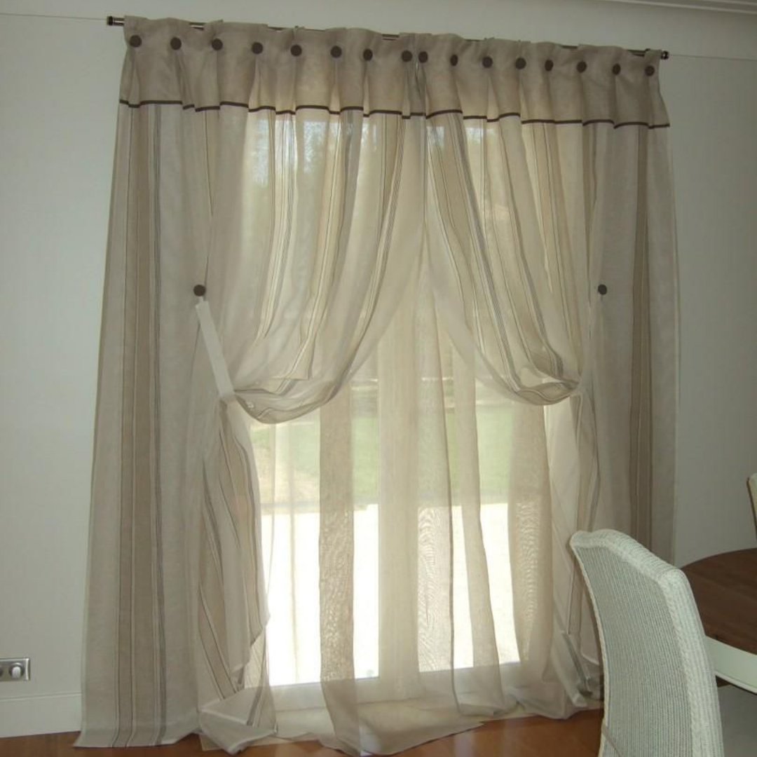 Des voilages beiges habillent une fenêtre. Les rideaux sont retenus par des embrasses.