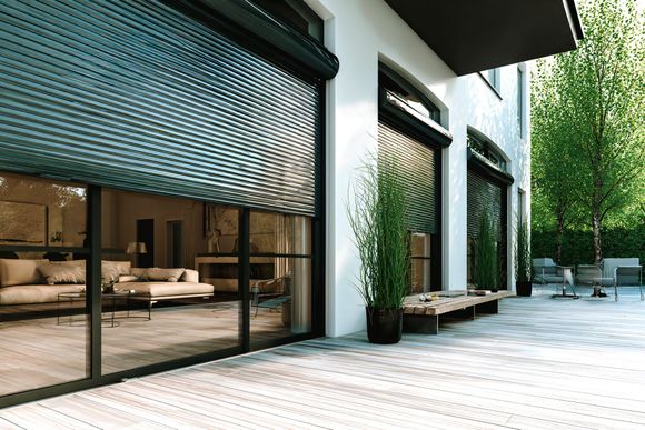 Patio moderne avec portes vitrées, stores tissés, murs blancs et arbres en pot le long d'une allée carrelée