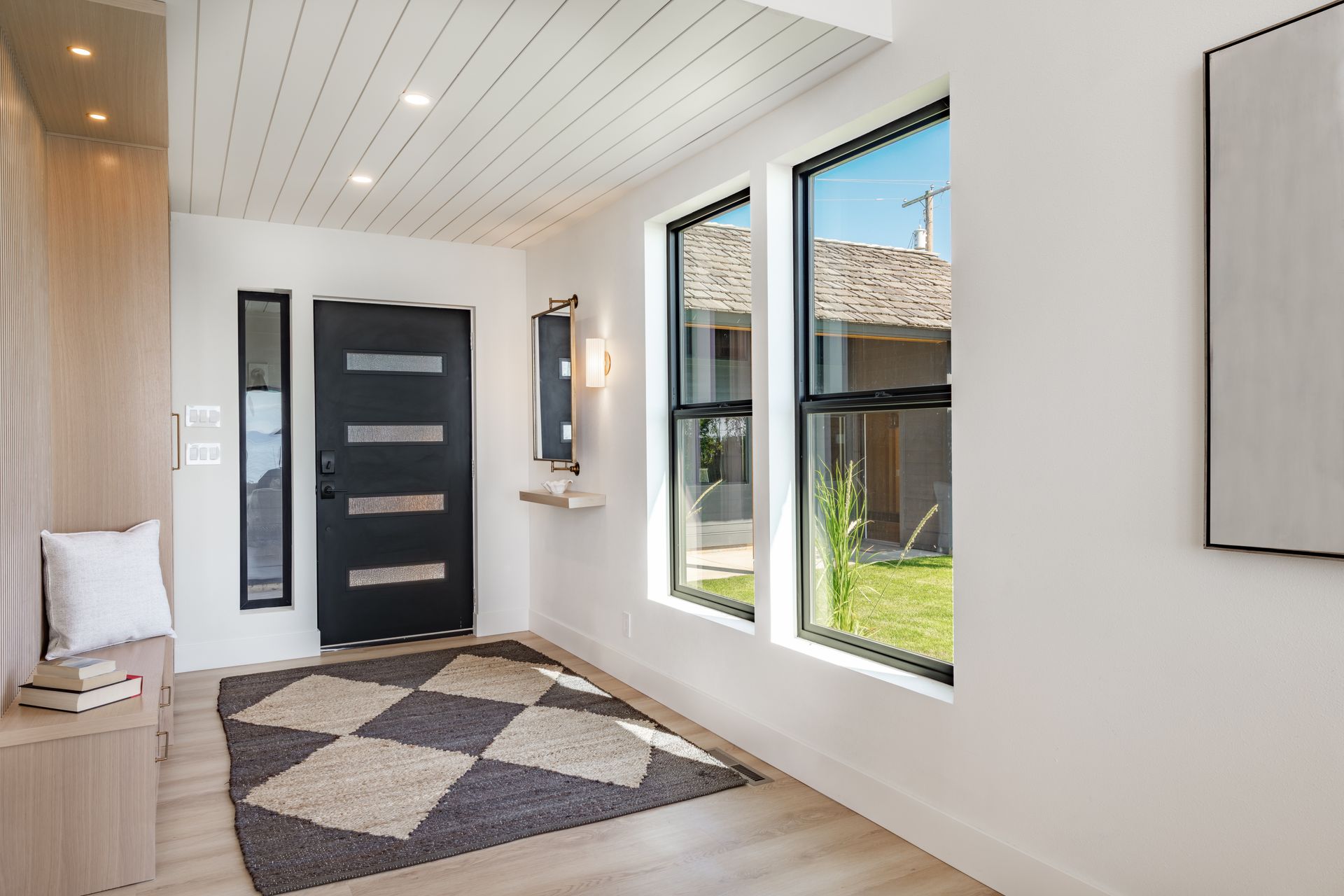 Entrée moderne avec porte d'entrée noire, tapis à motifs et grandes fenêtres donnant sur une cour