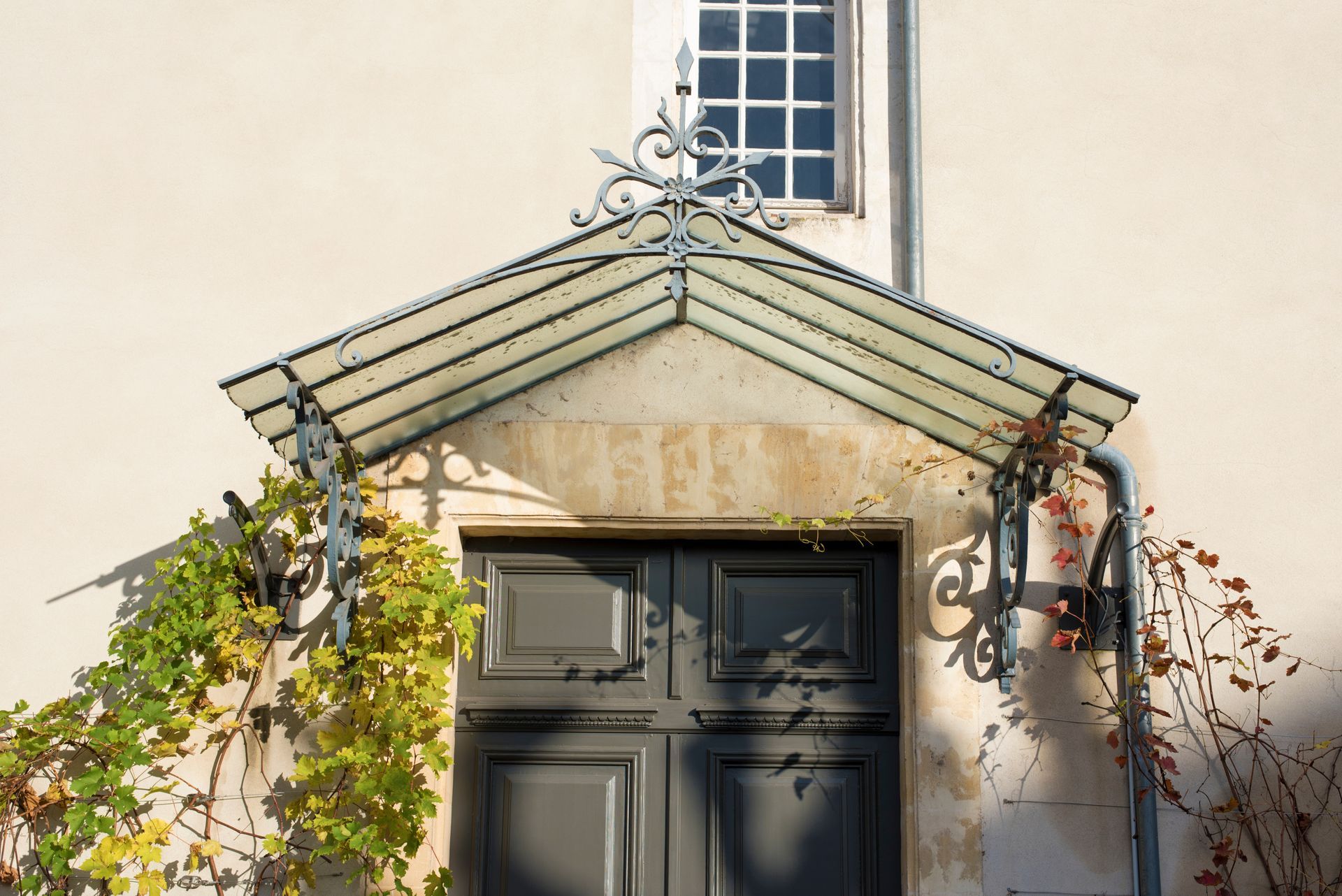 Marquise décorative en verre et en métal au-dessus d'une porte sombre sur un bâtiment couleur crème, avec des plantes grimpantes de part et d'autre.
