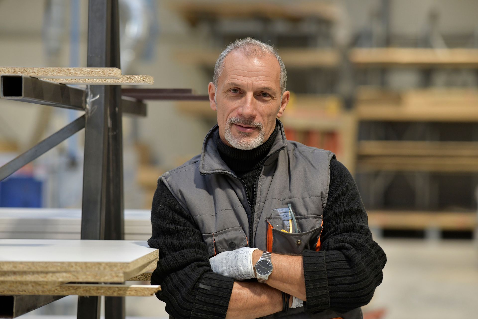 Une personne, les bras croisés, se tient debout dans un atelier, vêtue d'une veste noire et d'une montre, à côté d'étagères en bois empilées.