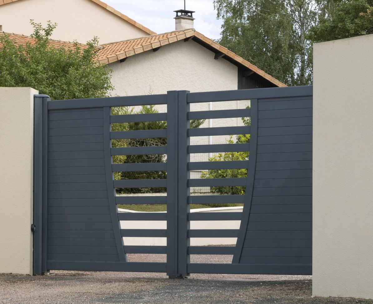 Portail d'allée moderne en métal gris entre des murs blancs, avec un design ajouré à claire-voie et des maisons visibles derrière.