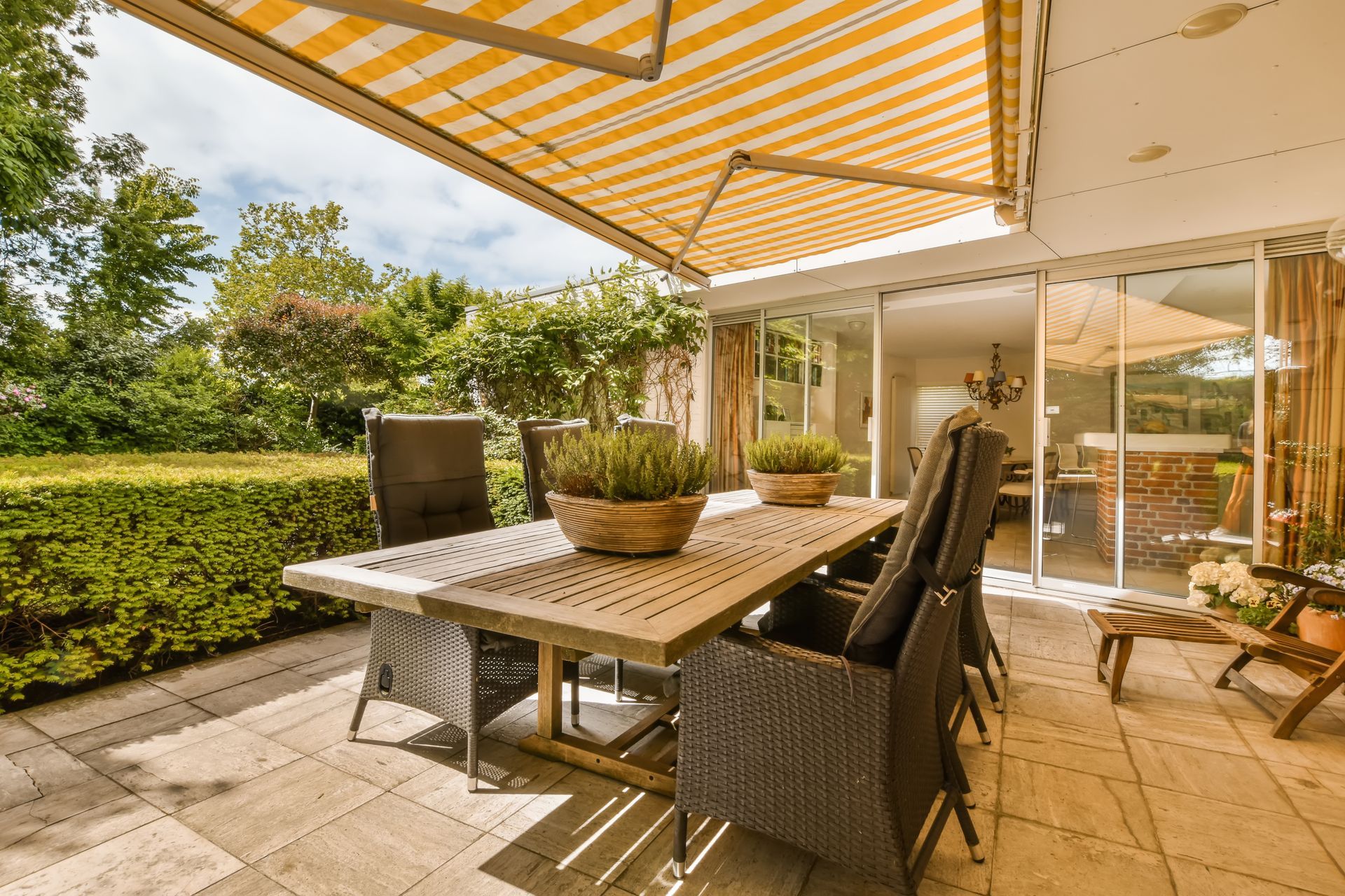 Terrasse avec salon de jardin en imitation rotin sous un store banne rayé, donnant sur un jardin verdoyant et des portes coulissantes en verre