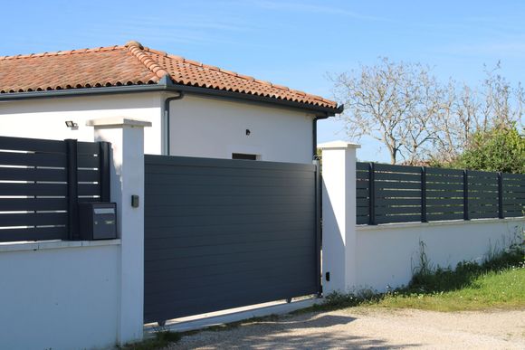 Maison blanche au toit de tuiles rouges, avec portail et clôture gris foncé, en bordure d'une route ensoleillée.