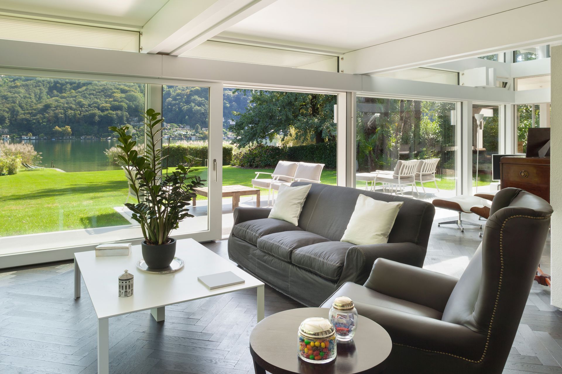 Véranda lumineuse avec canapés gris, table blanche, plante en pot et grandes fenêtres donnant sur un jardin verdoyant