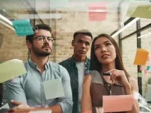 Tres colegas intercambian ideas mientras miran notas adhesivas en una pared de vidrio en un entorno de oficina.