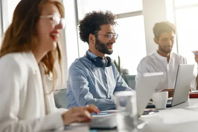 Tres personas sonriendo y trabajando con computadoras portátiles en una oficina luminosa; el hombre en el medio tiene auriculares.