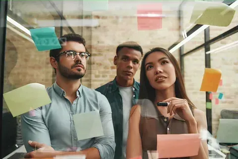 Tres colegas diversos intercambian ideas mientras miran notas adhesivas en una pared de vidrio en una oficina.