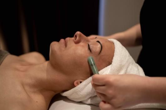 Une personne recevant un soin du visage au pinceau, allongée sur une table de spa et enveloppée dans une serviette blanche.