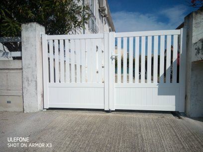 Un portail en métal blanc à lattes verticales se dresse entre deux piliers de pierre sur une allée en béton.