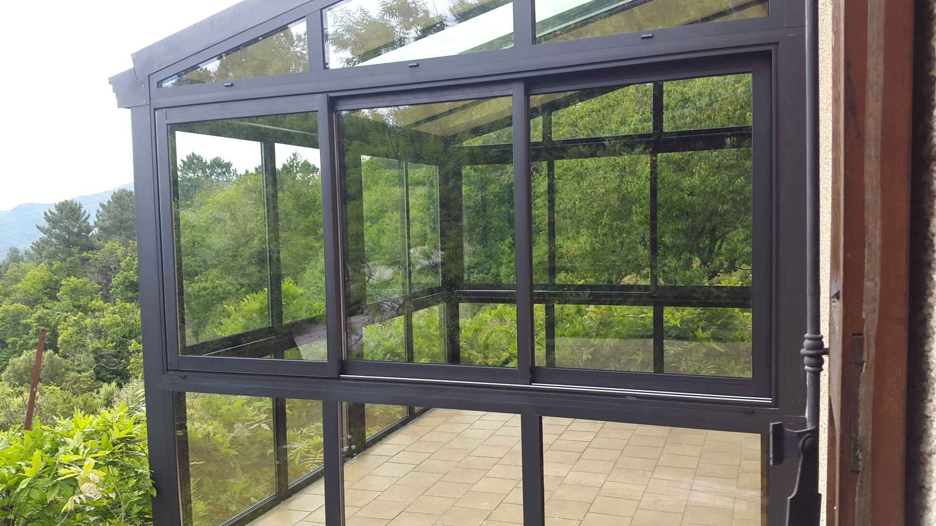 Une véranda ou un jardin d'hiver avec une structure en métal sombre et de grandes baies vitrées, donnant sur une forêt verdoyante luxuriante.