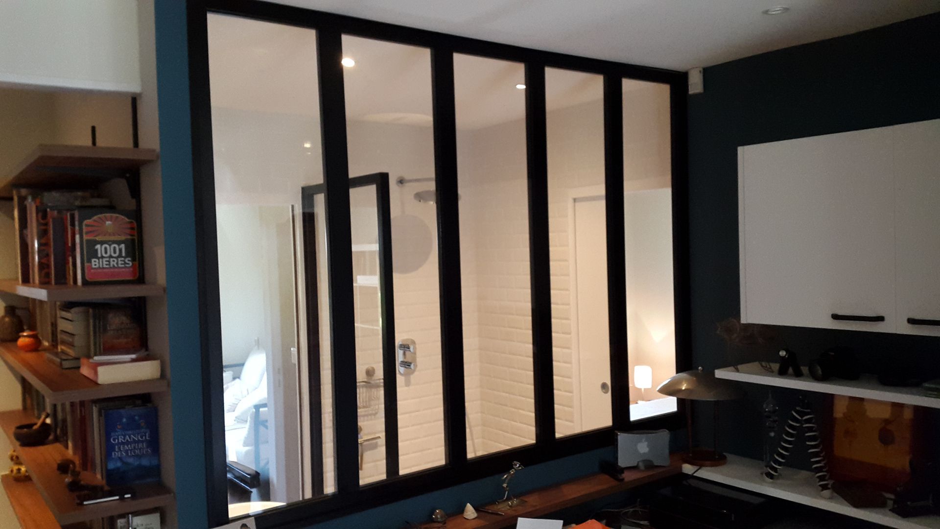 Une cloison en verre de style industriel, à cadre noir, séparant un espace de vie d'une salle de bains aux murs carrelés de blanc.